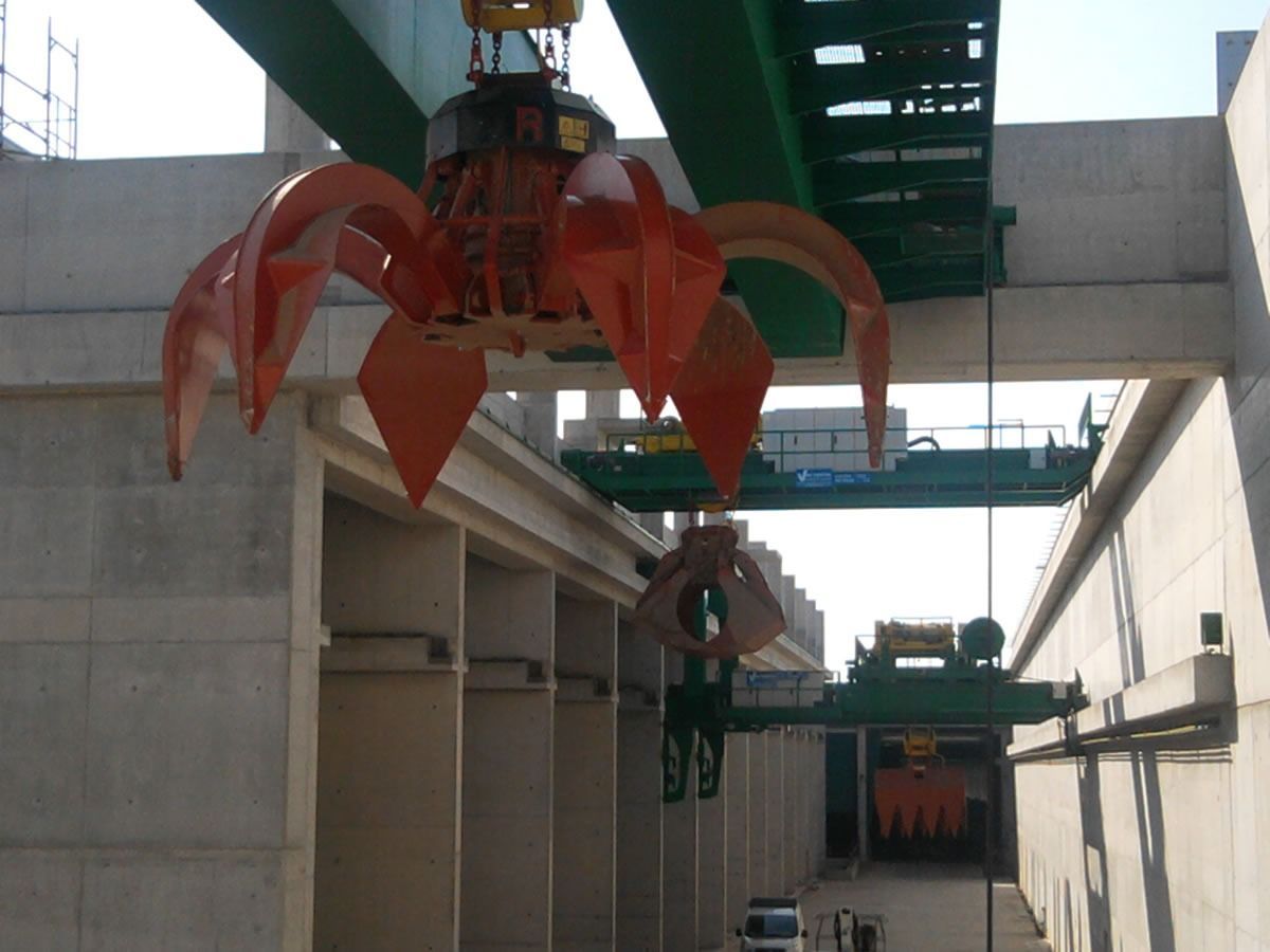 Gru a ponte con pinza arancione per il sollevamento, installata in una struttura in cemento.
