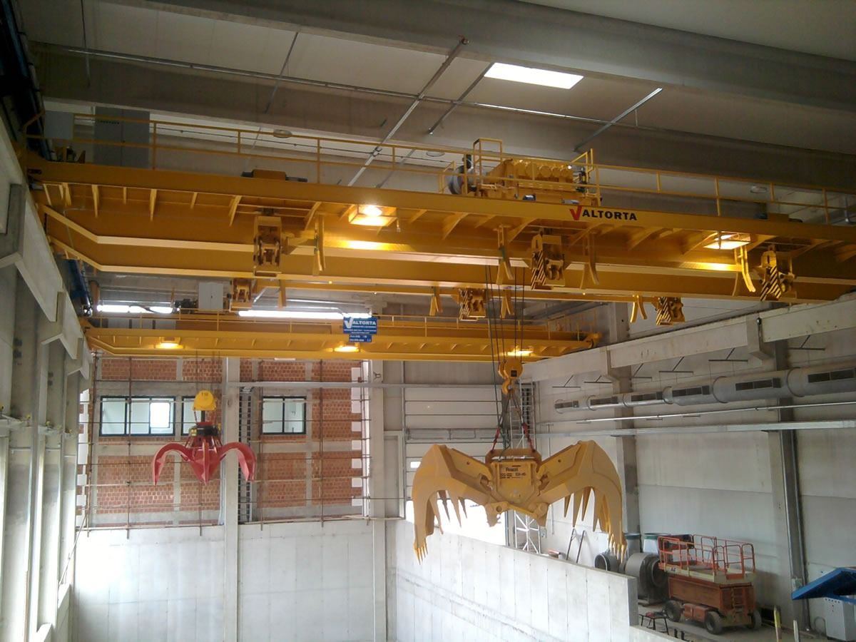 Gru a ponte gialla con pinza aperta su una fossa di cemento, probabilmente in un ambiente industriale.