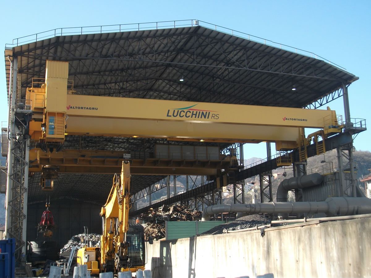 Gru a ponte gialla all'interno di un impianto di trattamento dei rifiuti. Un escavatore carica il materiale.