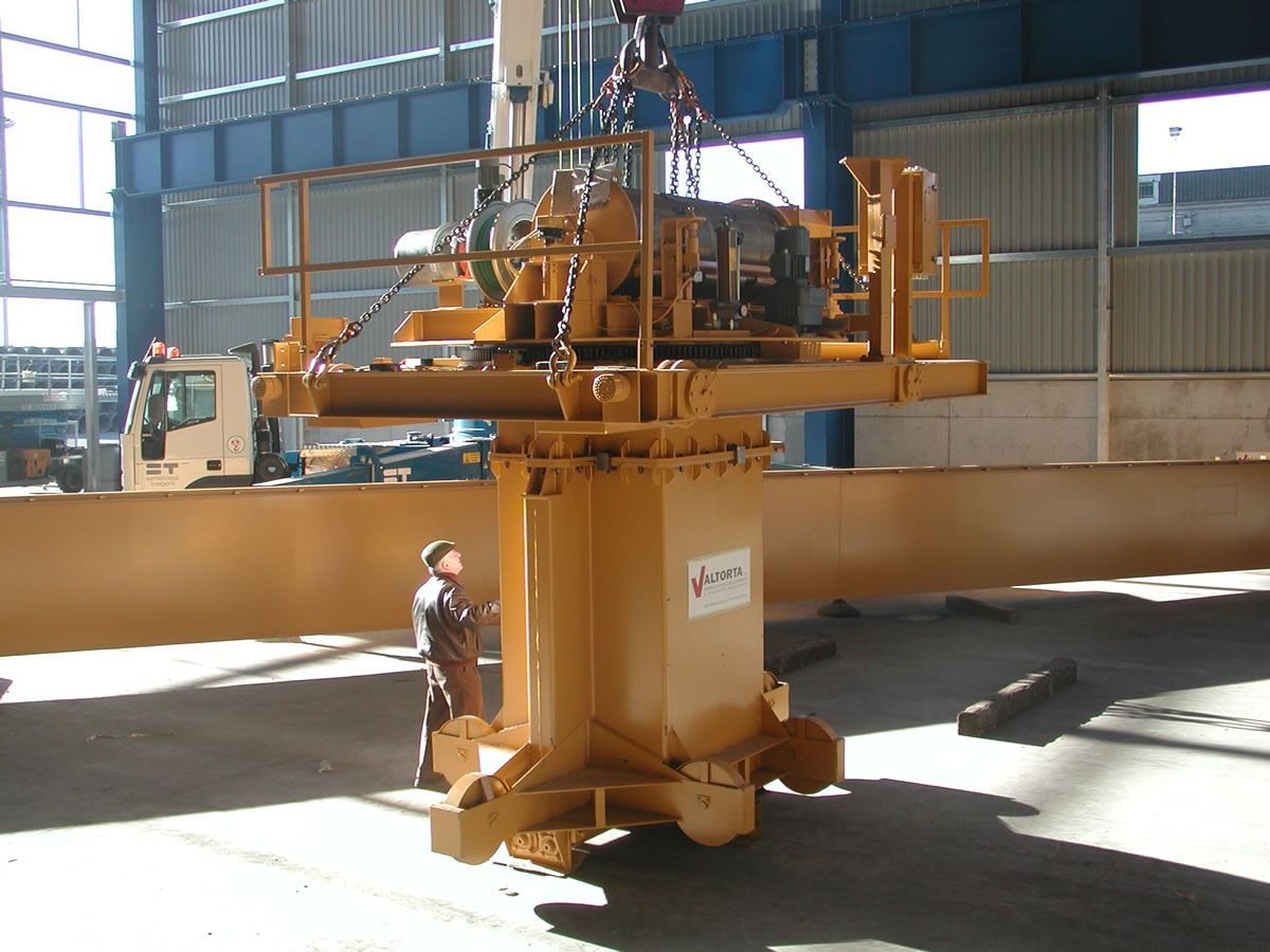 Macchina industriale gialla sollevata da una gru; un operaio è in piedi lì vicino in un grande magazzino.