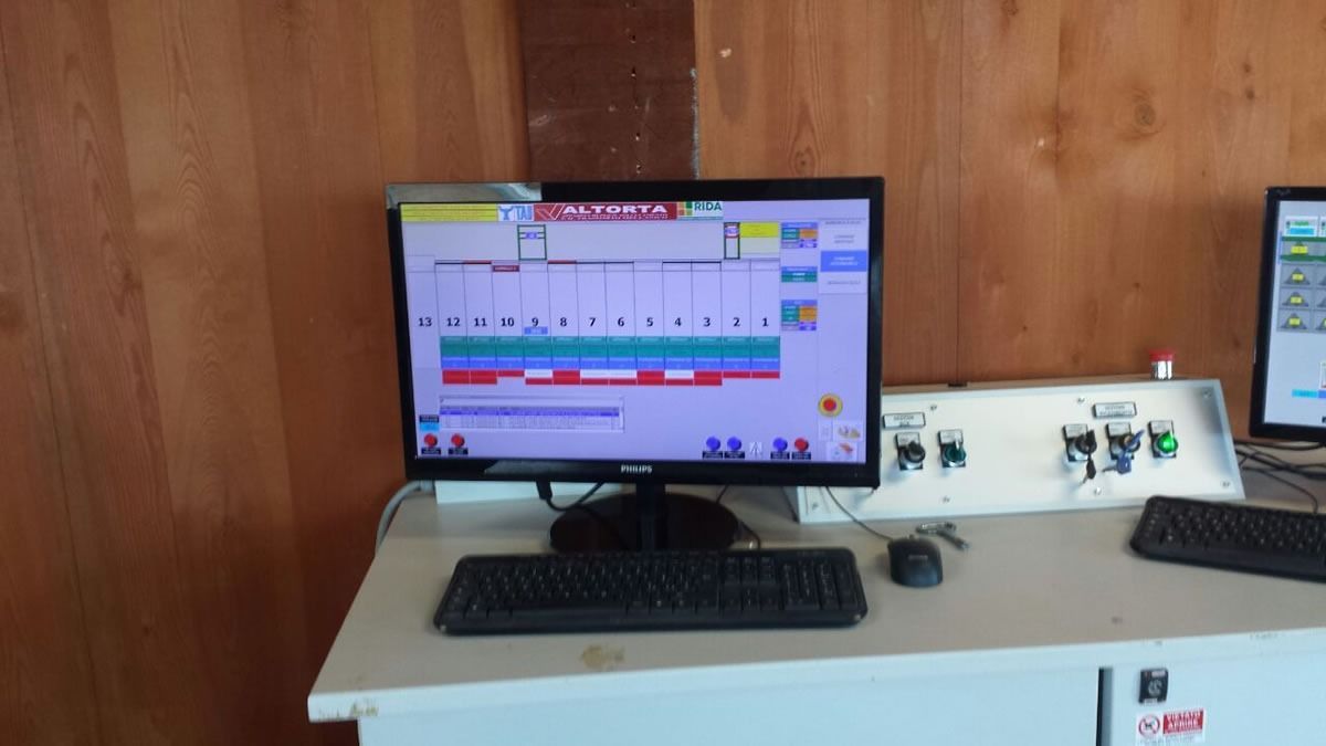 Monitor del computer che visualizza dati, tastiera, mouse e pannello di controllo su una scrivania bianca con pannelli in legno.