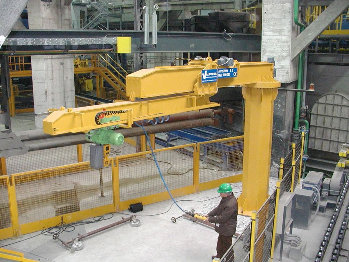 Gru industriale gialla che solleva un tubo metallico; operaio che aziona la gru in una fabbrica.