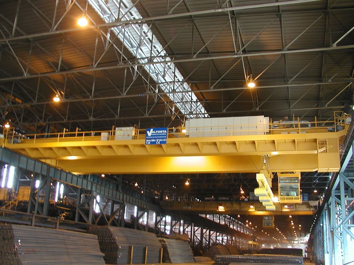 Gru a ponte gialla in un magazzino, intenta a sollevare materiali. Sono visibili travi metalliche e pile di articoli.