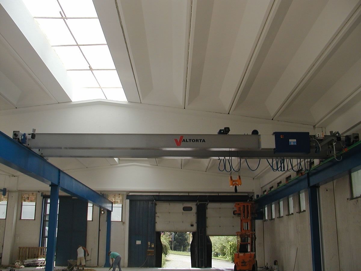 Gru a ponte in un magazzino, con struttura in acciaio blu e grigio. Sotto la gru c'è un ascensore.