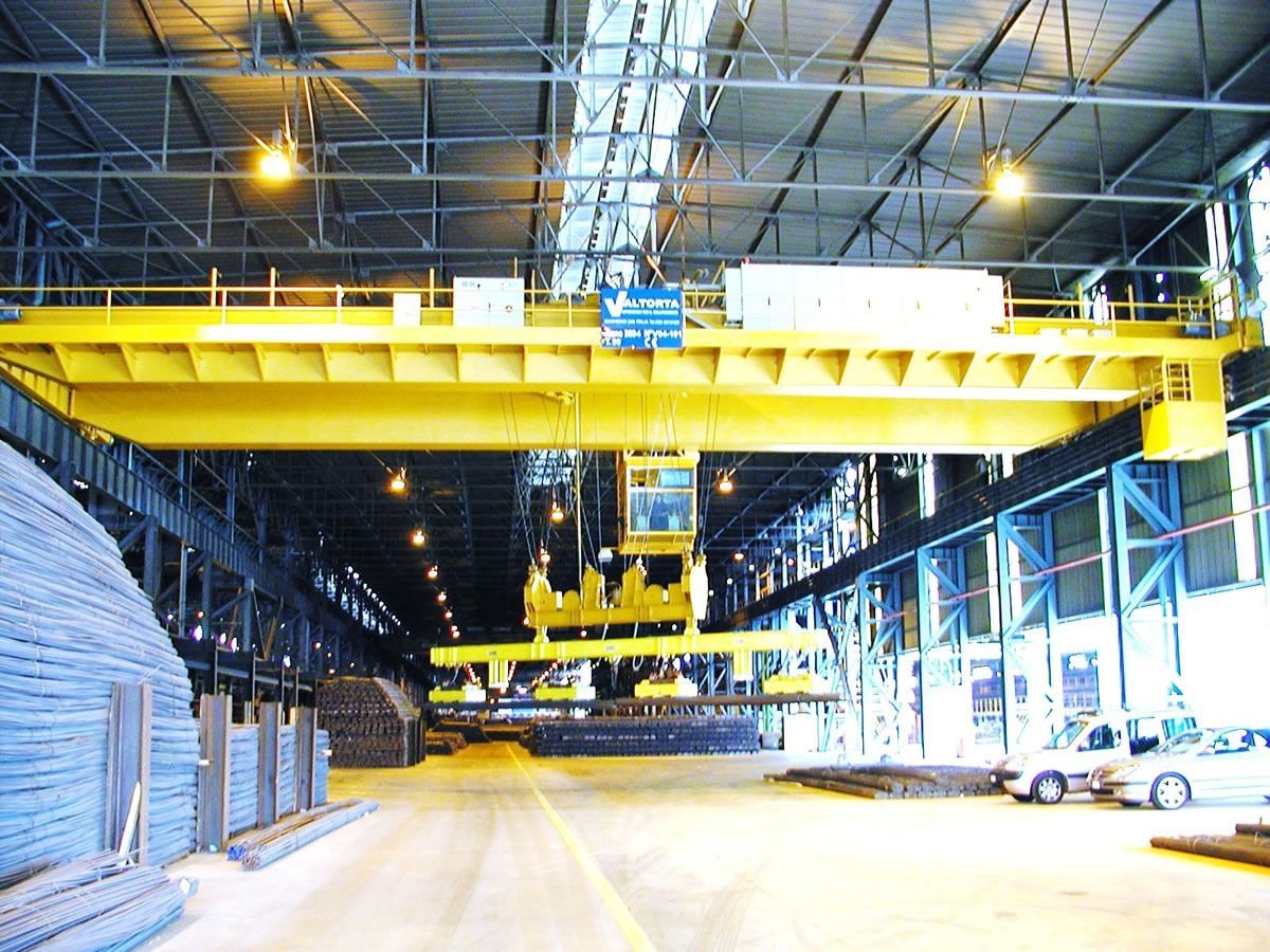 Gru a ponte gialla che solleva travi d'acciaio all'interno di un grande magazzino.