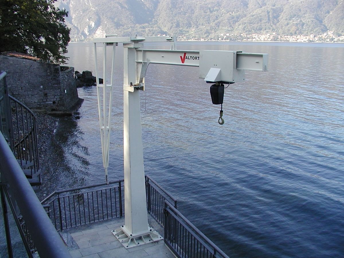 Gru bianca protesa sull'acqua, montata su una piattaforma di cemento con ringhiera, sullo sfondo lago e montagne.