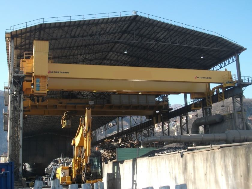 Una gru a ponte gialla all'interno di un magazzino. Un escavatore lavora sotto.