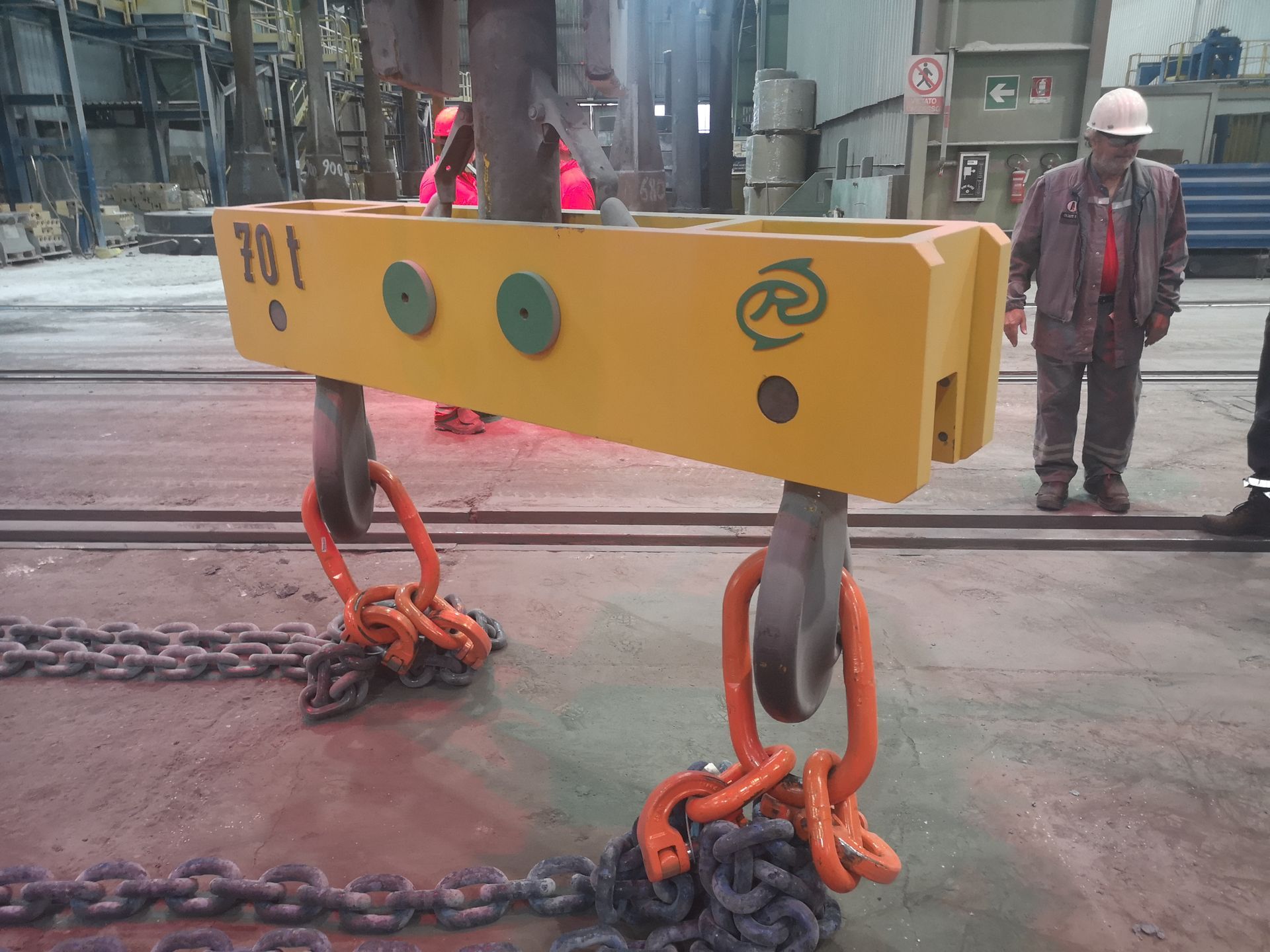 Trave di sollevamento gialla con ganci e catene arancioni, appoggiata sul pavimento. Un operaio è in piedi nelle vicinanze.