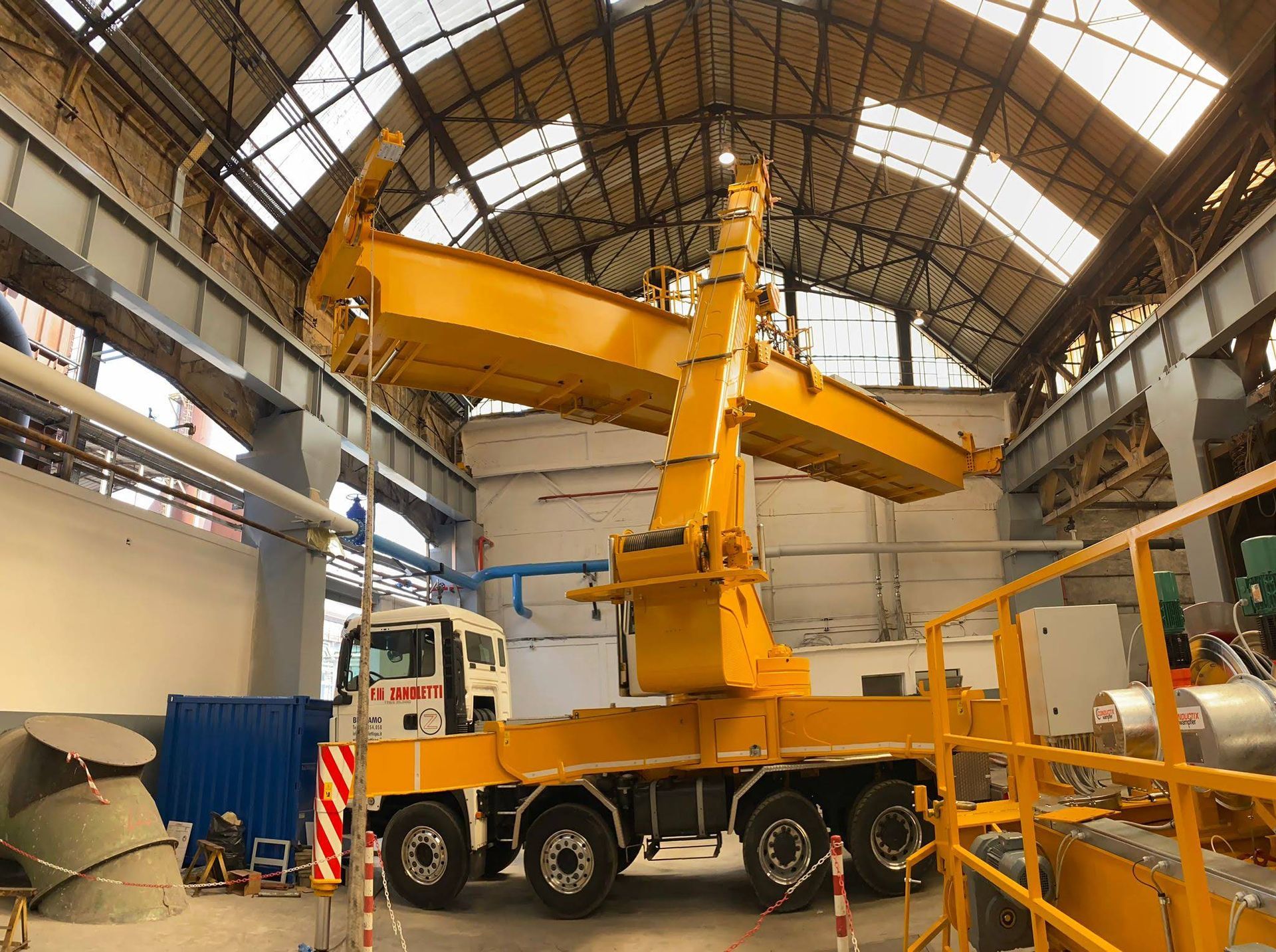 Gru industriale gialla che solleva una grande trave gialla all'interno di un magazzino. È visibile un camion bianco.