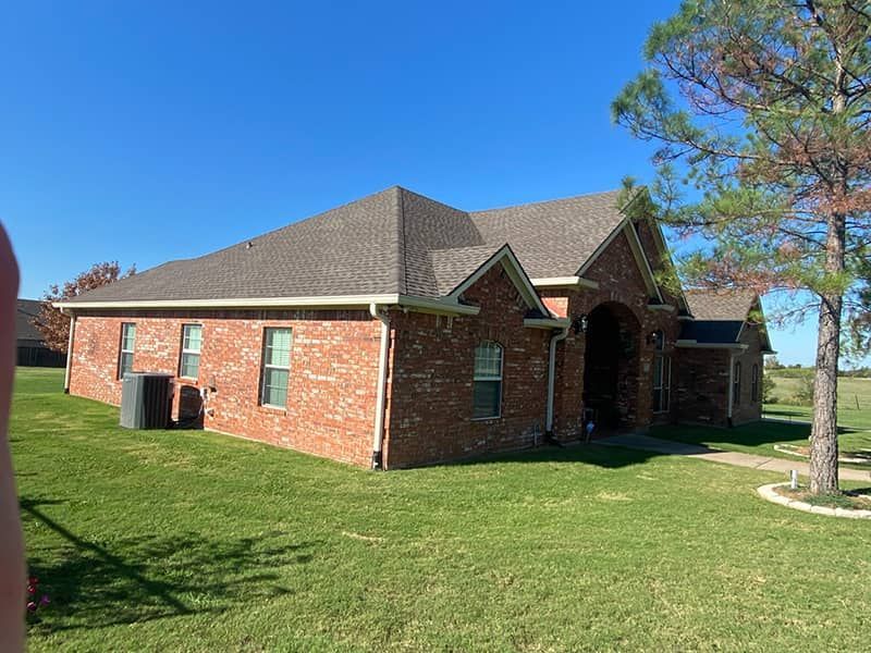A brick house with a gray roof is sitting on top of a lush green field.