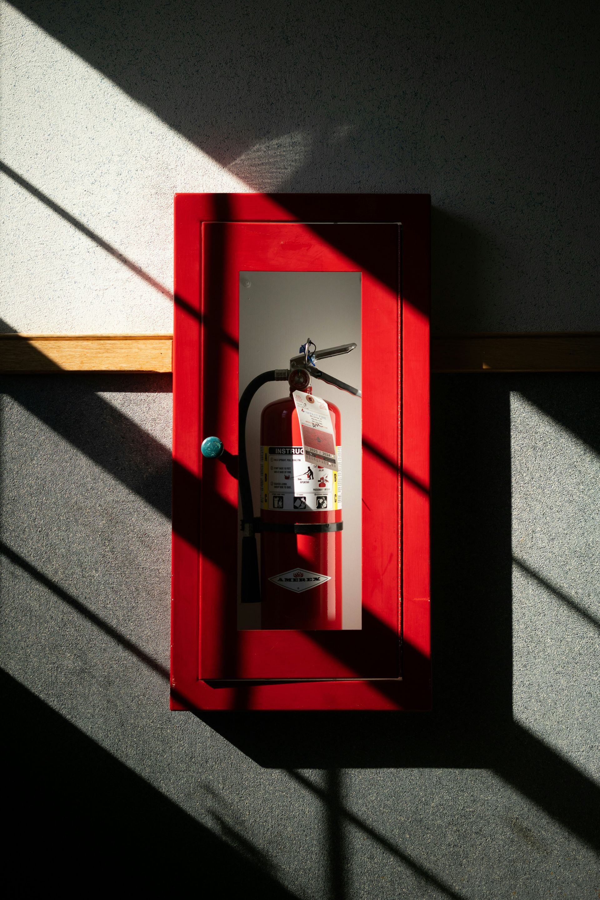 Armoire à extincteurs rouge sur un mur texturé, partiellement éclairée par la lumière du soleil.