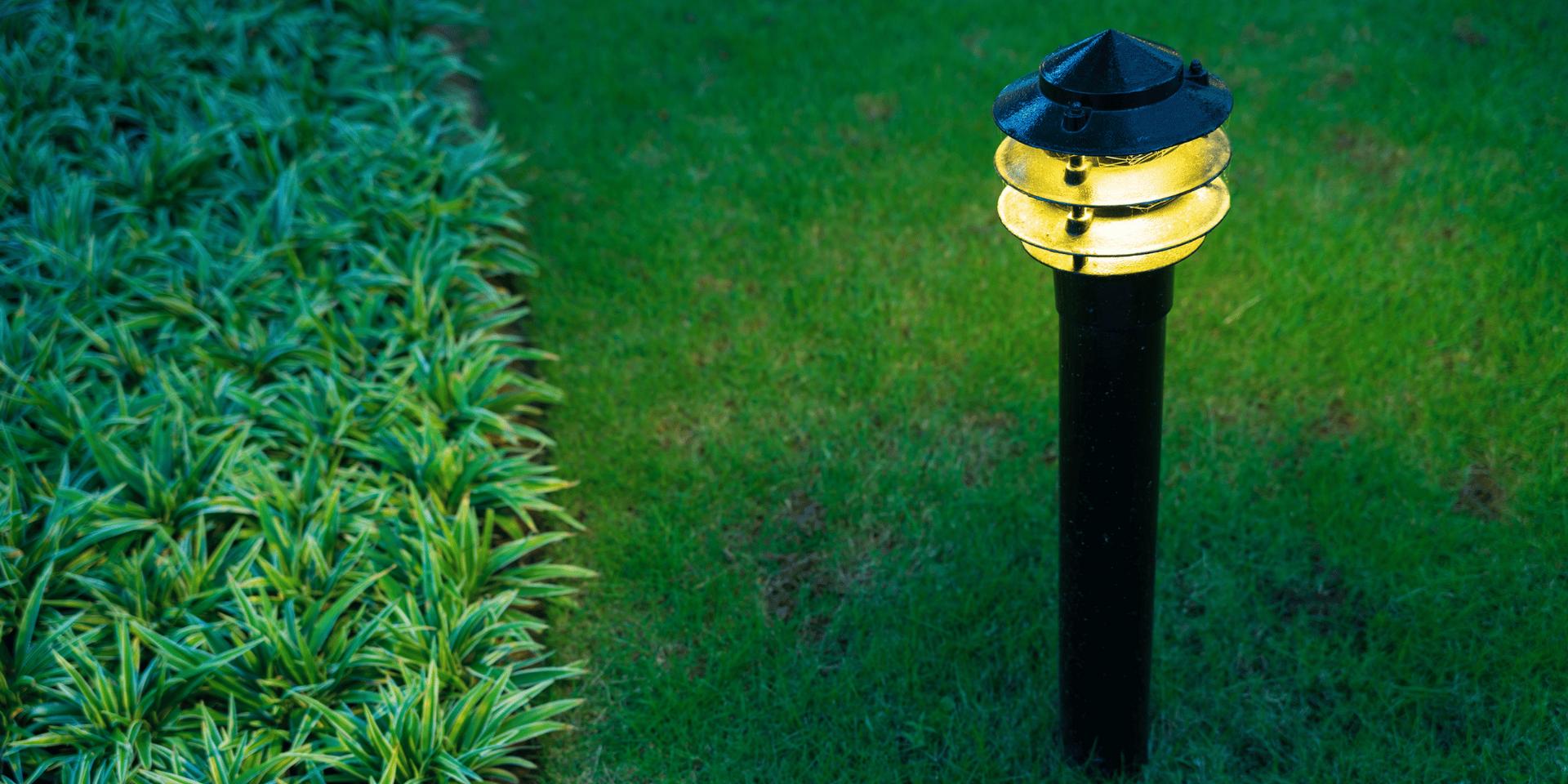 Lit garden lamp on green grass, next to a bed of leafy green plants.