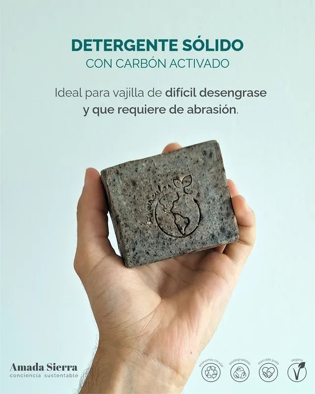 Una mano sostiene una pastilla de detergente sólido color carbón. Texto: «Detergente sólido con carbón activado».