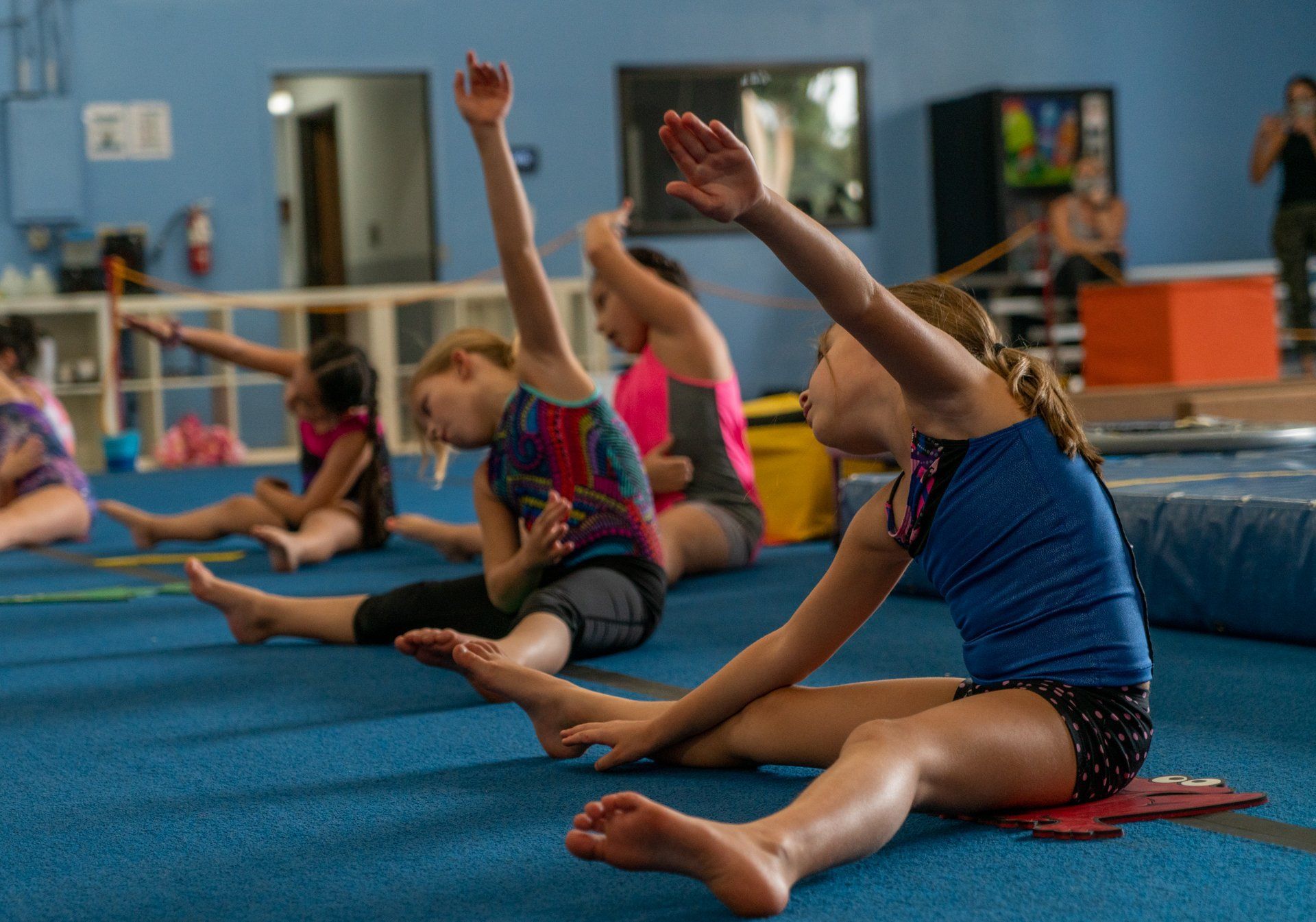 A group of young girls are stretching on the floor in a gym.