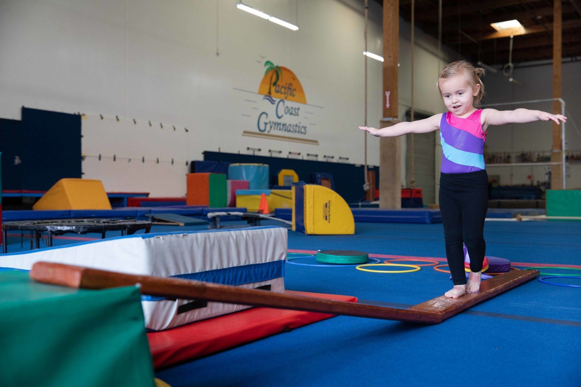 A little girl is balancing on a wooden balance beam in a gym.