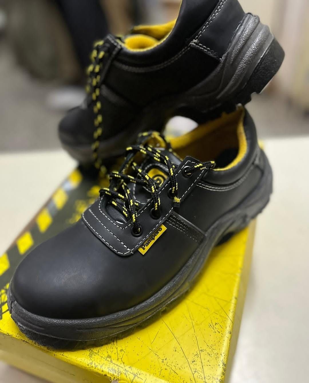 Botas de trabajo negras con costuras y cordones amarillos sobre una caja amarilla, mostradas en un entorno comercial.