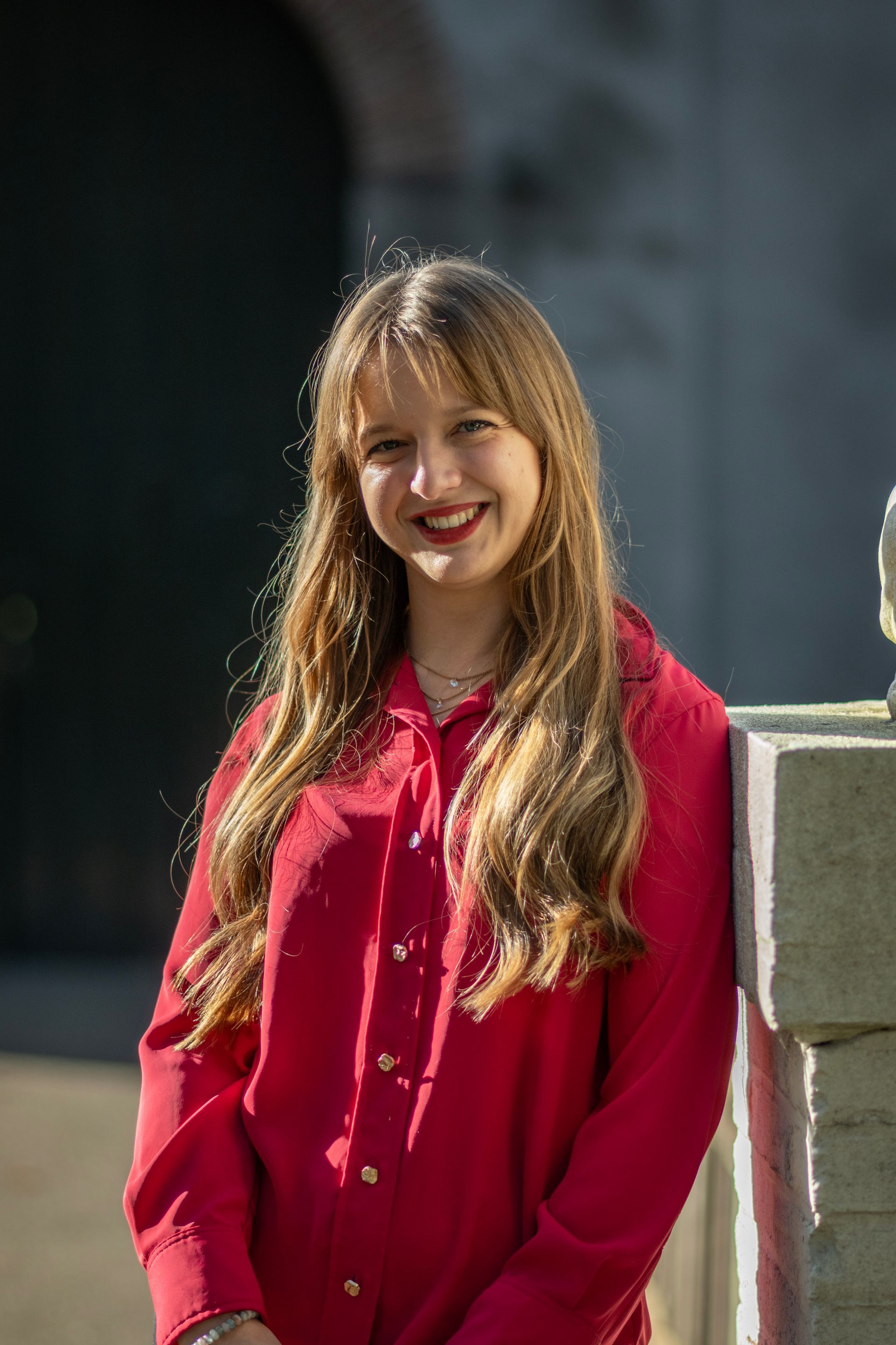 Een vrouw in een rode blouse en zwarte broek zit glimlachend op een beige bank.