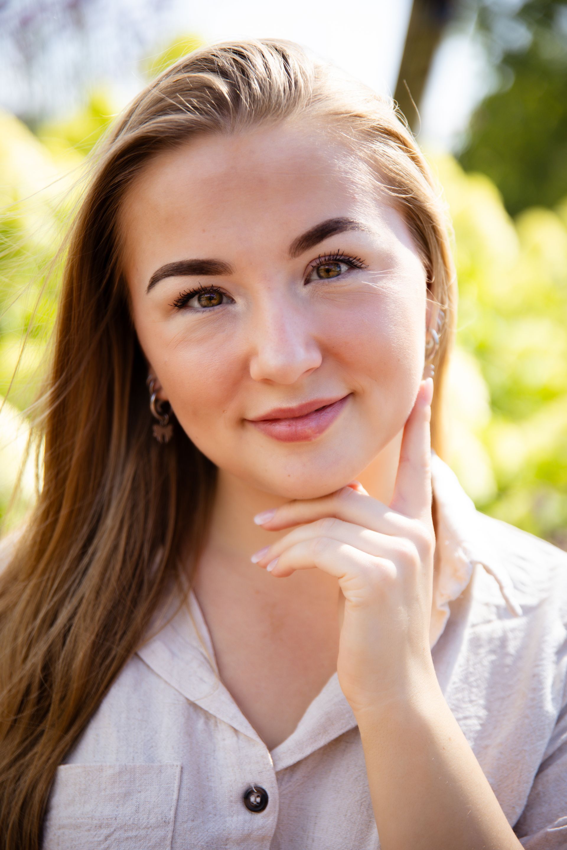 Vrouw met lang blond haar lacht, raakt haar kin aan en poseert buiten.