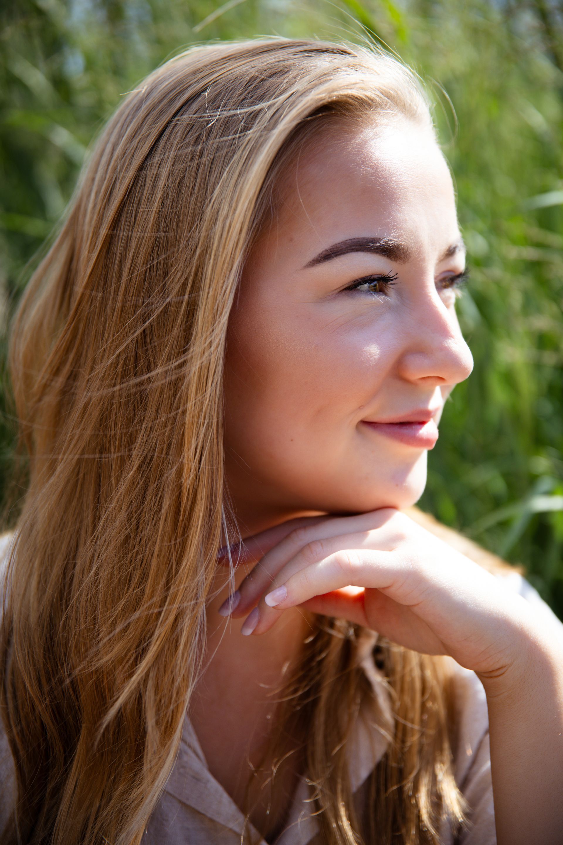 Blonde vrouw rust met haar kin op haar hand en lacht in het zonlicht, buiten met op de achtergrond groen gebladerte.