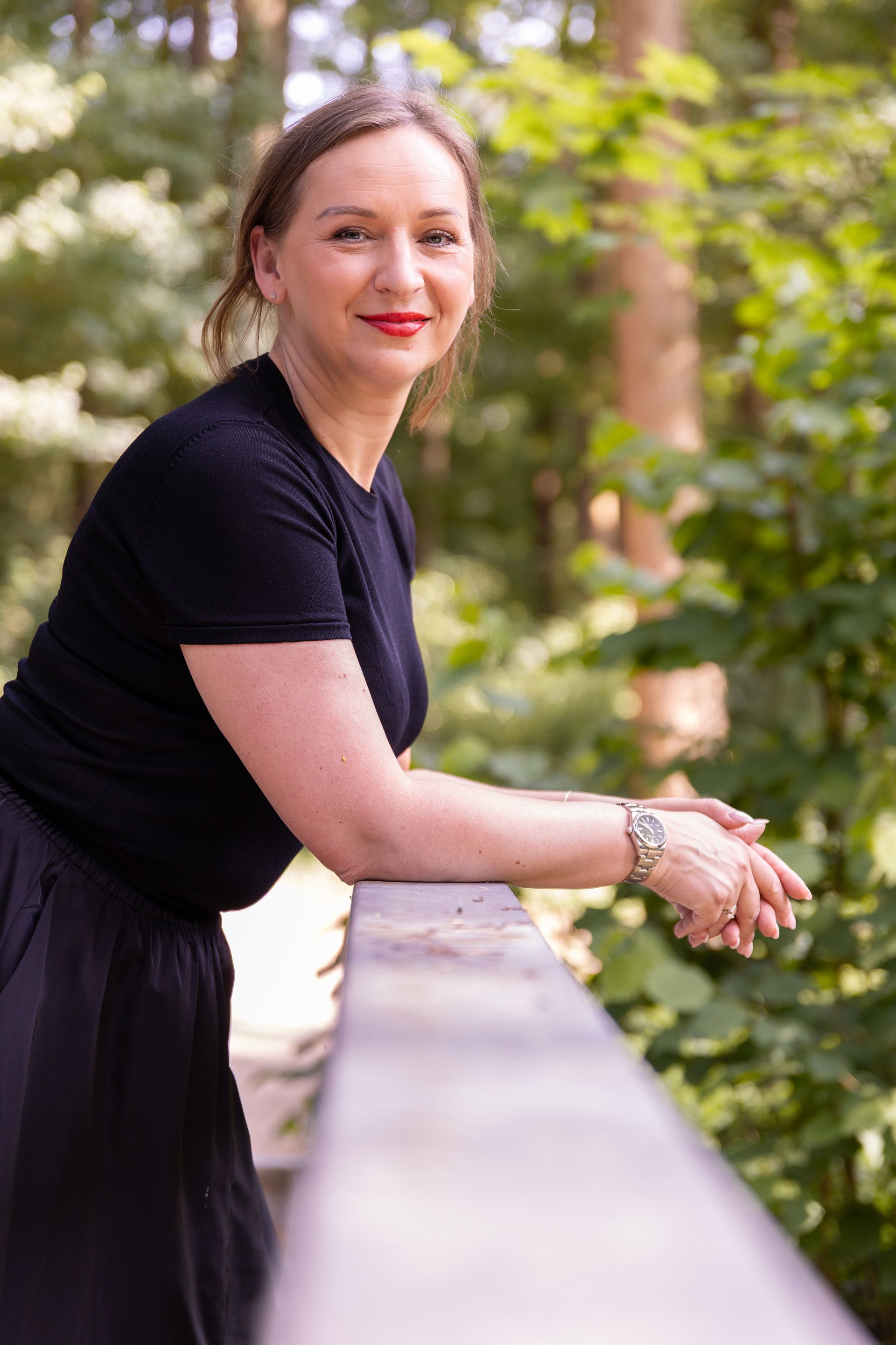 Vrouw leunend tegen een houten balustrade, glimlachend. Gekleed in een zwarte top, rok en lippenstift, buiten.