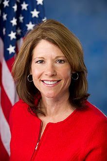A woman in a red jacket is smiling in front of an american flag.