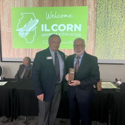 Two men in suits and ties are standing next to each other in front of a sign that says welcome il corn growers association.