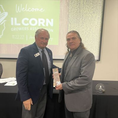 Two men are standing in front of a sign that says welcome il corn growers association
