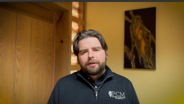 A man with a beard is standing in front of a painting on a wall.