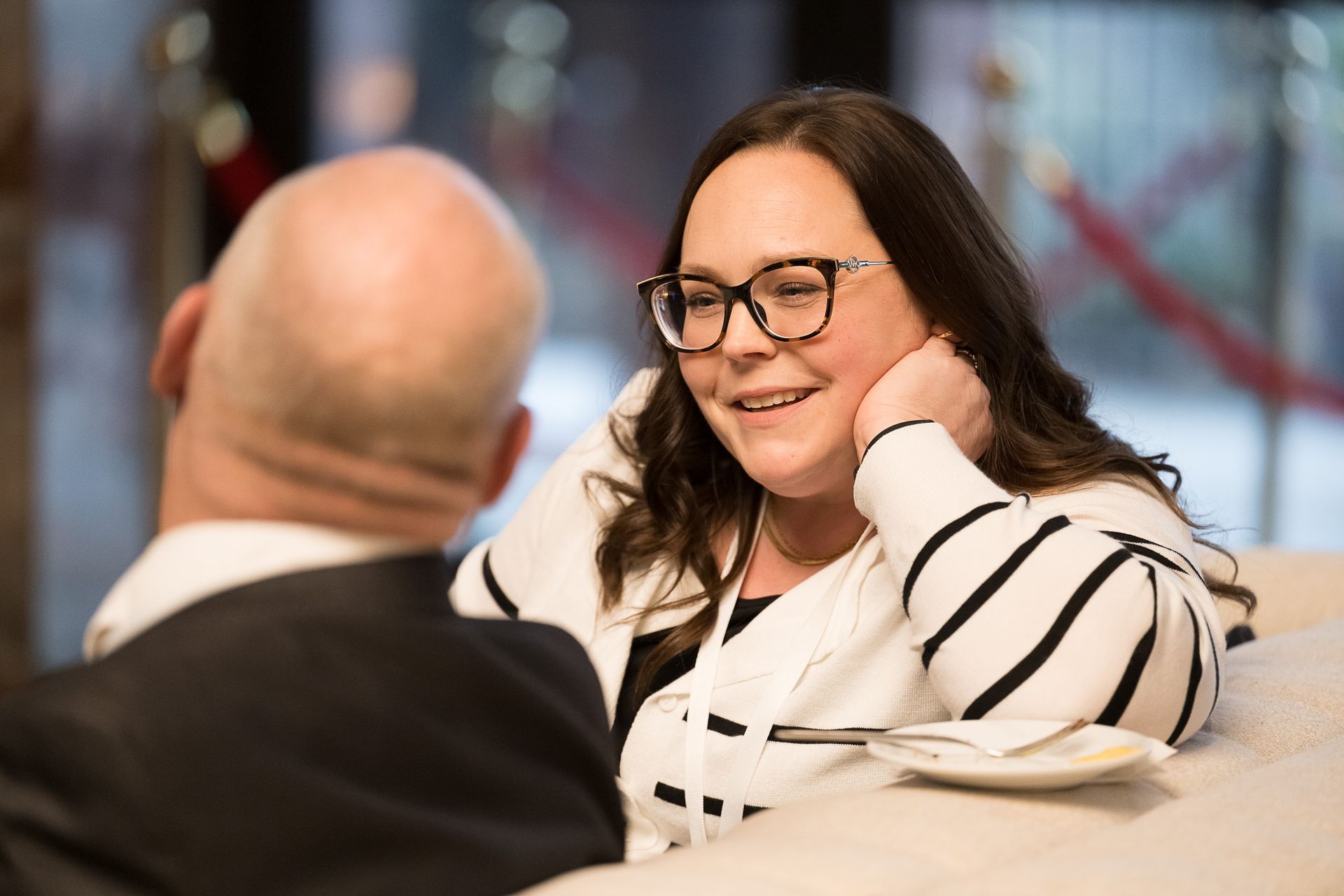 Woman in glasses smiles, resting hand on cheek, talking with a person, seated indoors.