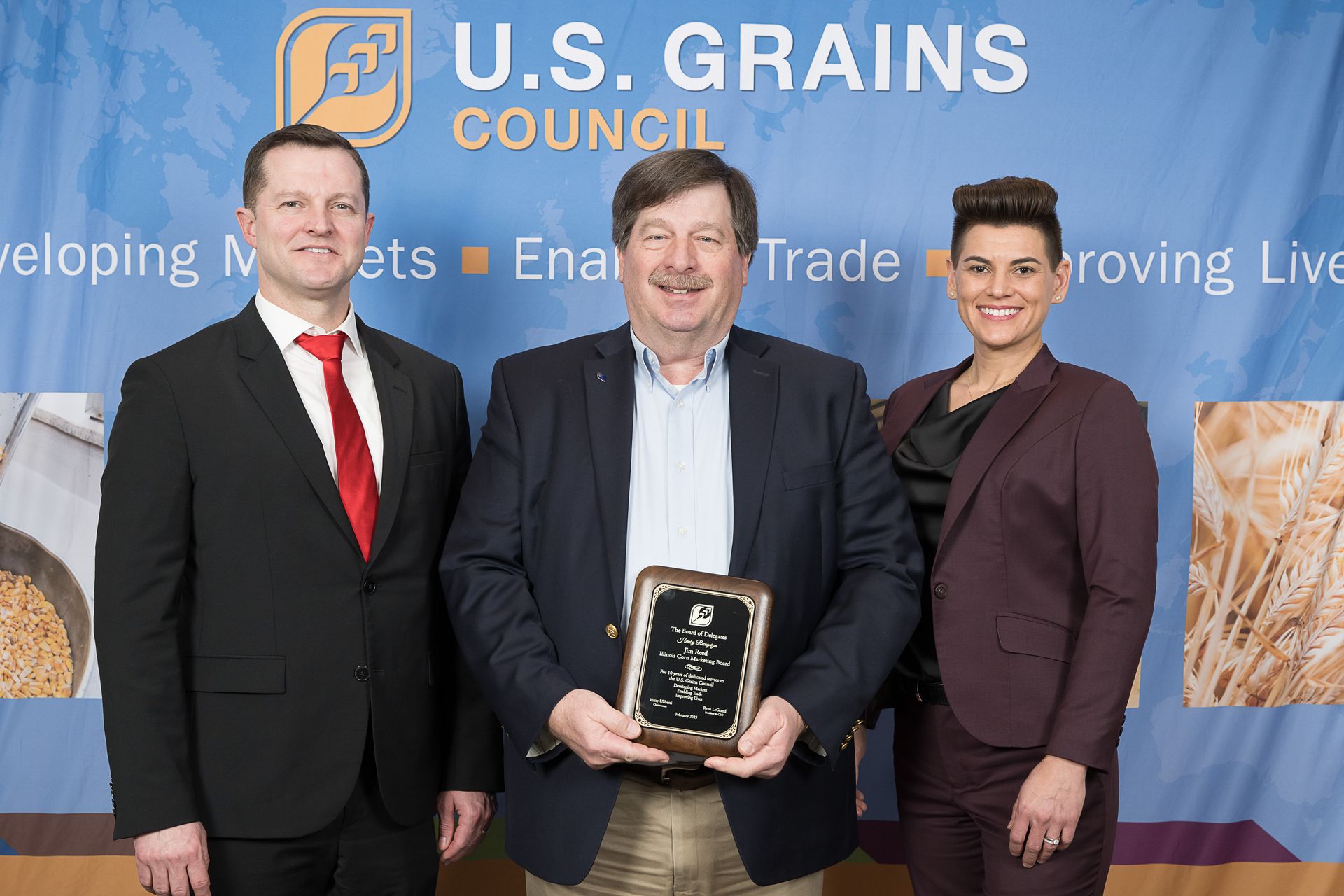 Three people at a podium with a US Grains Council backdrop. A man holds an award; all are smiling.