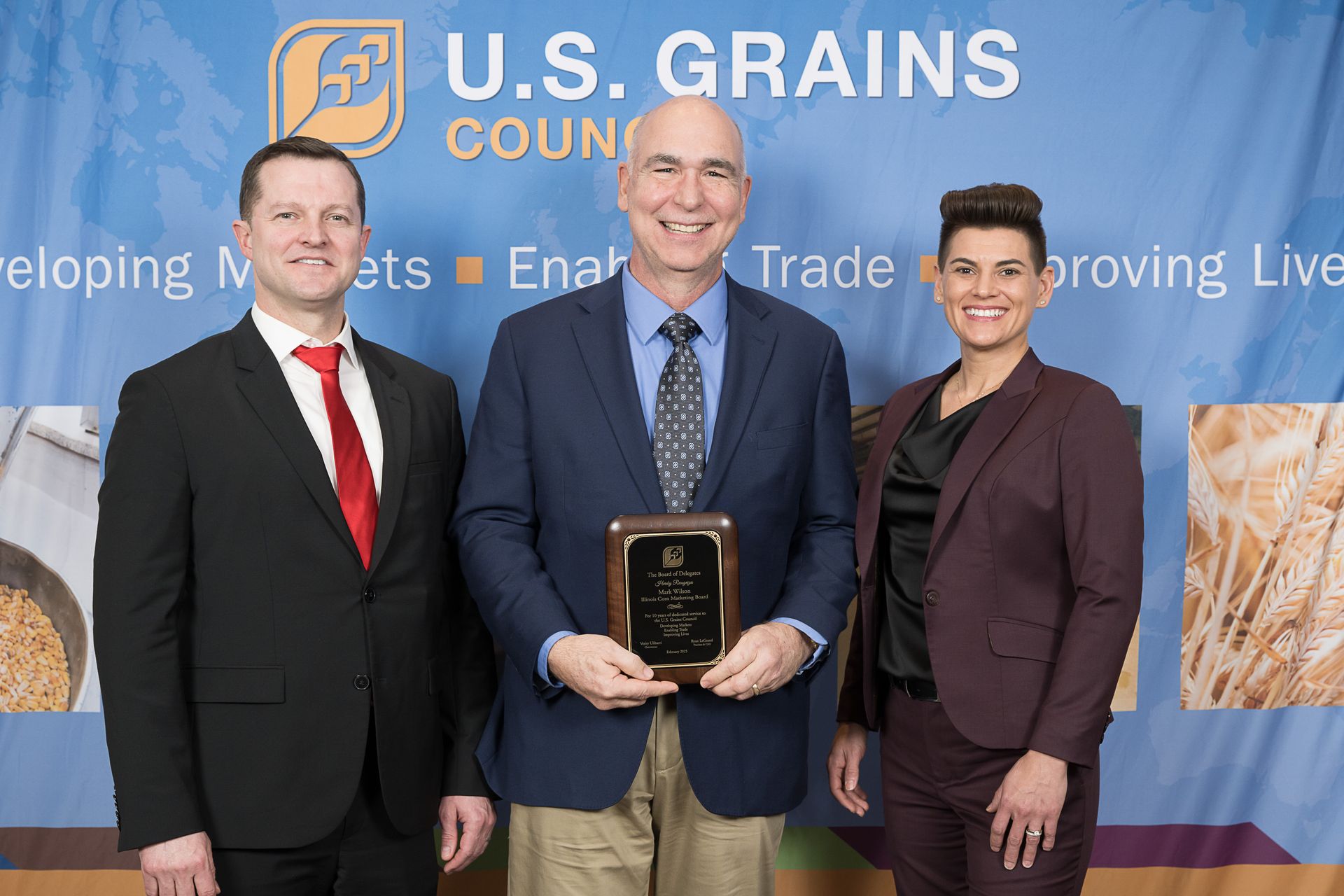 Three people at US Grains Council event: man holding plaque, flanked by two others. Background banner.