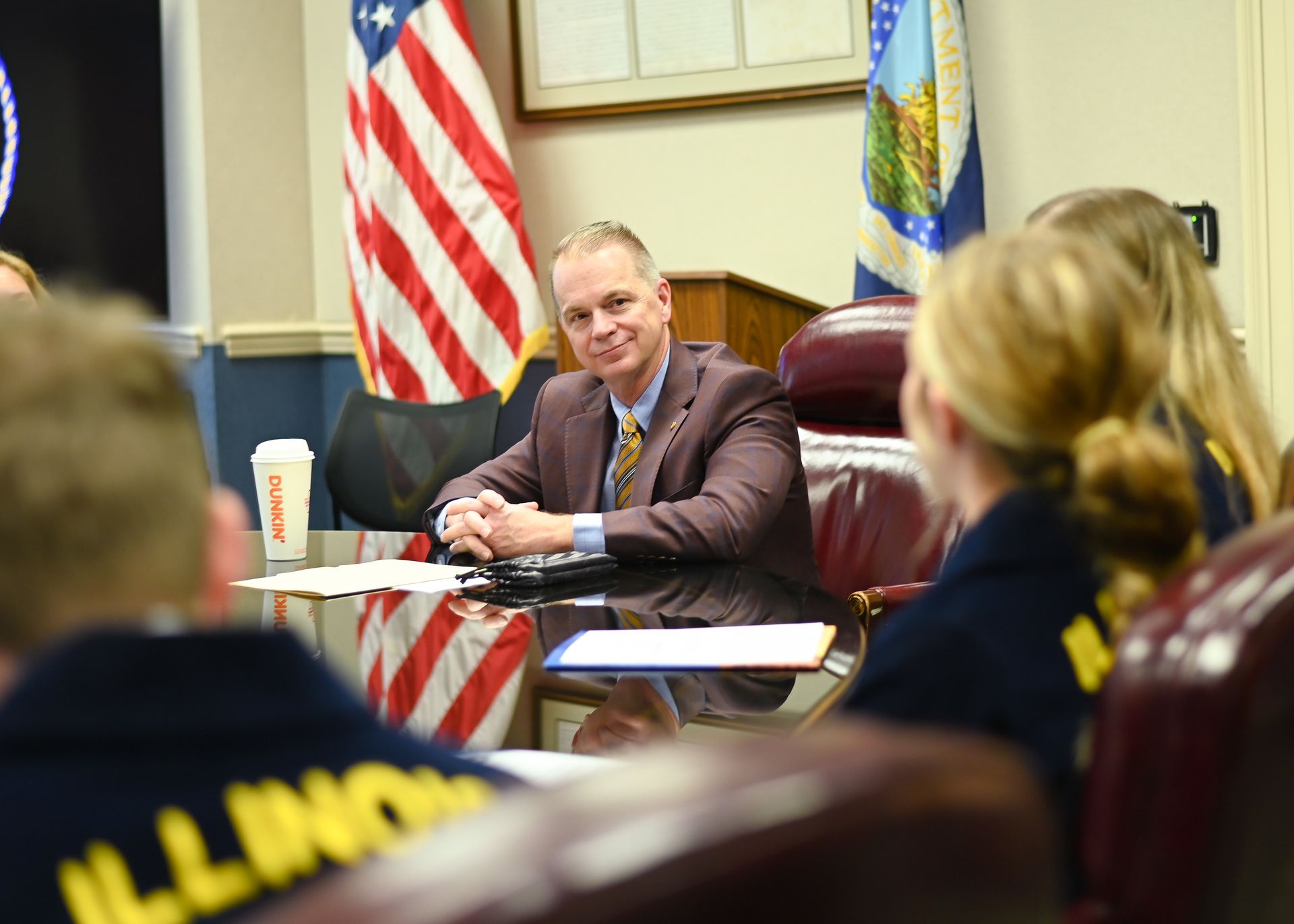 Man in suit smiles at a table with people in blue shirts, US and state flags in background.