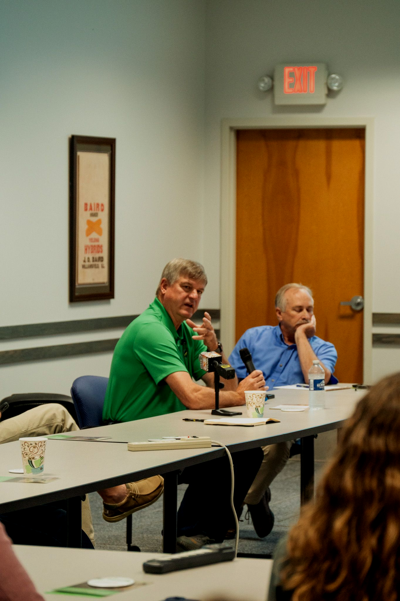 Two men seated at a table, speaking. One in green, gesturing; the other in blue, listening. Indoor office setting.