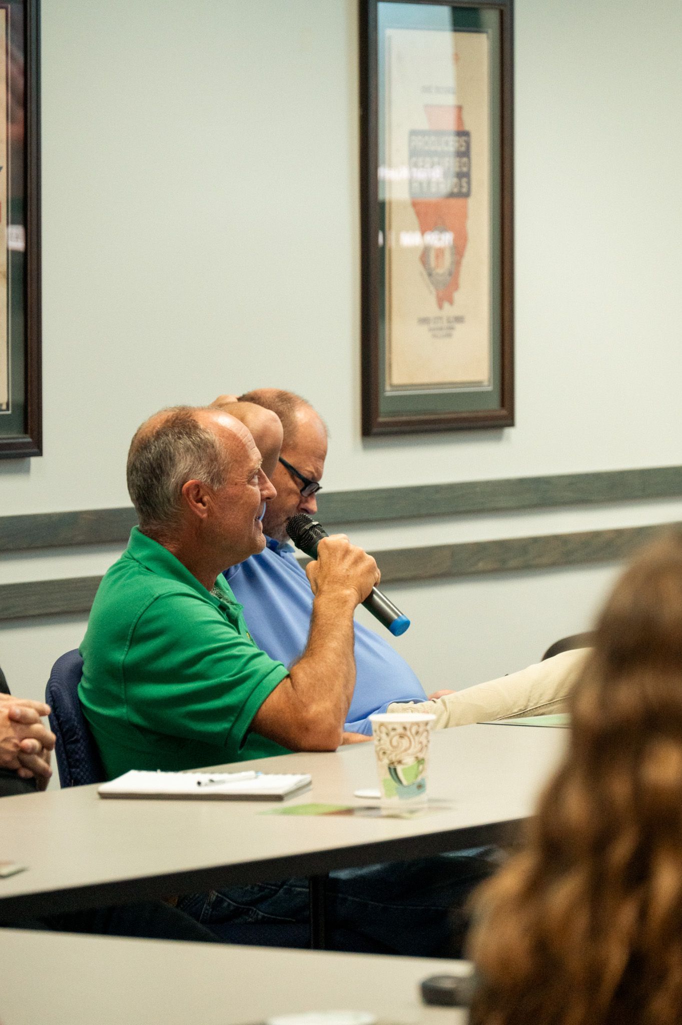 Man in green shirt speaks into microphone, sitting at a table with another man.