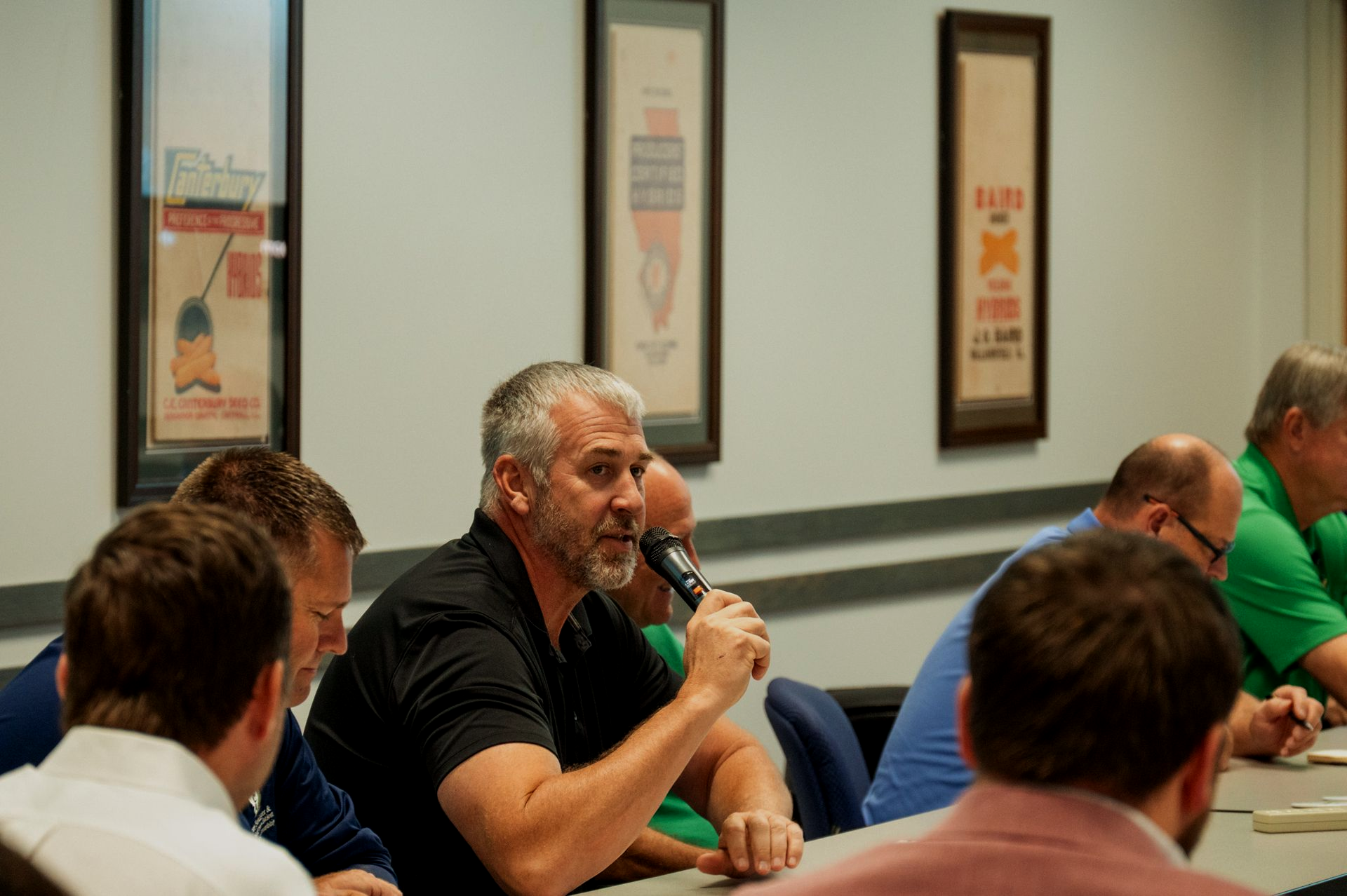 Man speaking into microphone at a table. Other people seated around him in a conference room.
