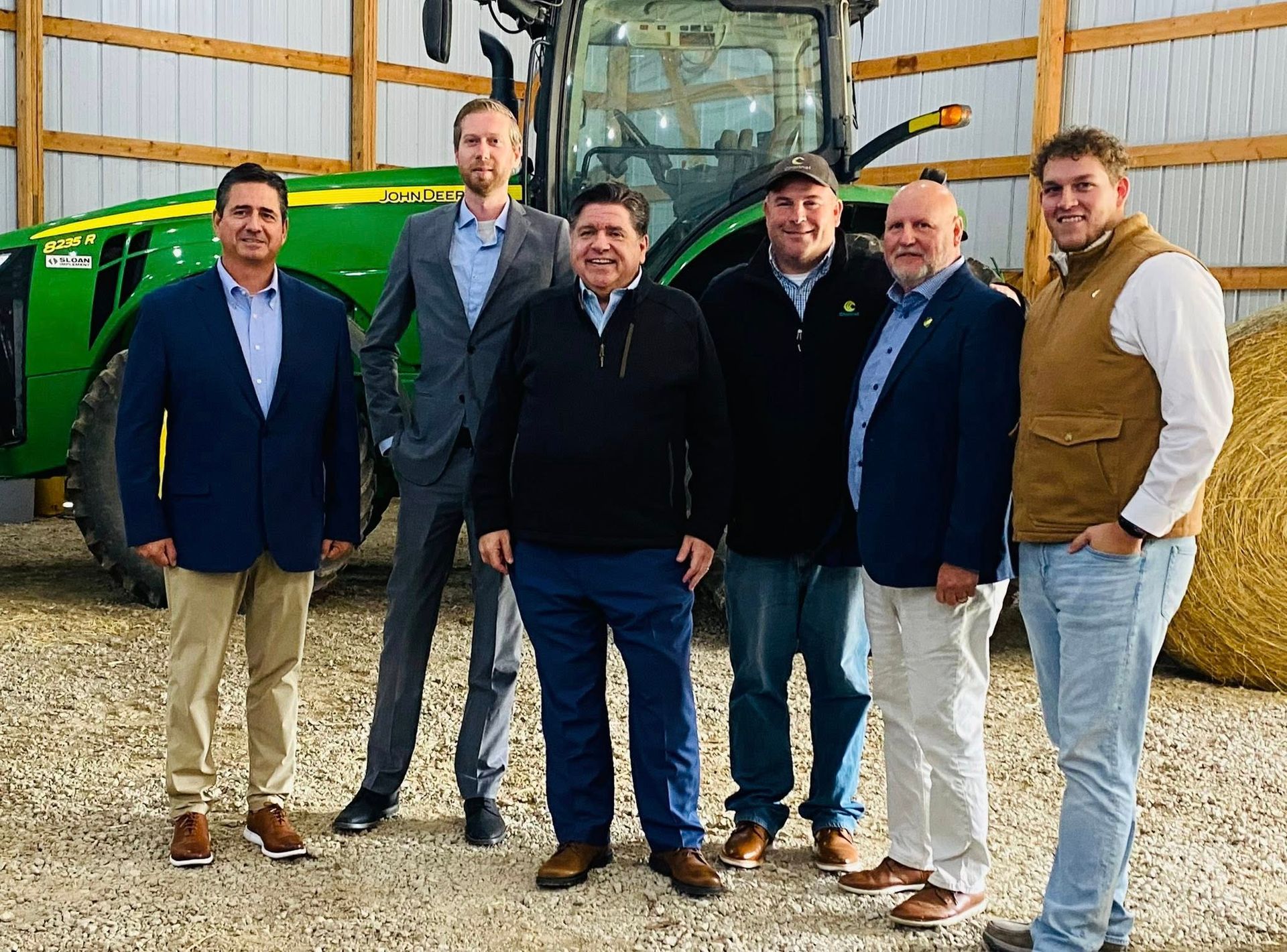 Six men in business attire stand in a barn near a tractor.