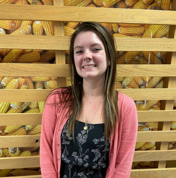 A woman in a pink cardigan is smiling in front of a wall of corn