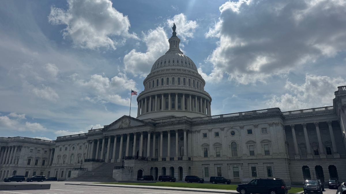 The U.S. Capitol Building with its white dome, columns, and flag against a partly cloudy blue sky.