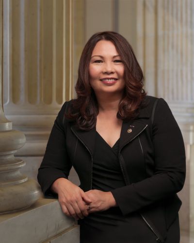 A woman in a black jacket and dress is leaning against a pillar.