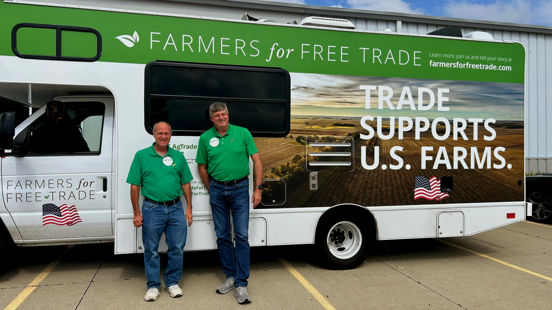 Two men in green shirts stand in front of a bus with “Farmers for Free Trade” and “Trade Supports U.S. Farms” slogans.