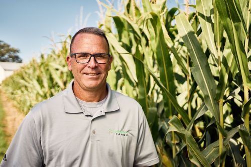 A man wearing glasses is standing in front of a corn field.