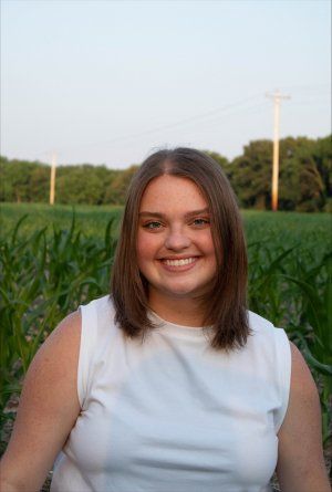 A young woman in a white tank top is smiling in front of a corn field.