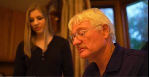 A man with glasses is sitting at a table with a woman standing behind him.