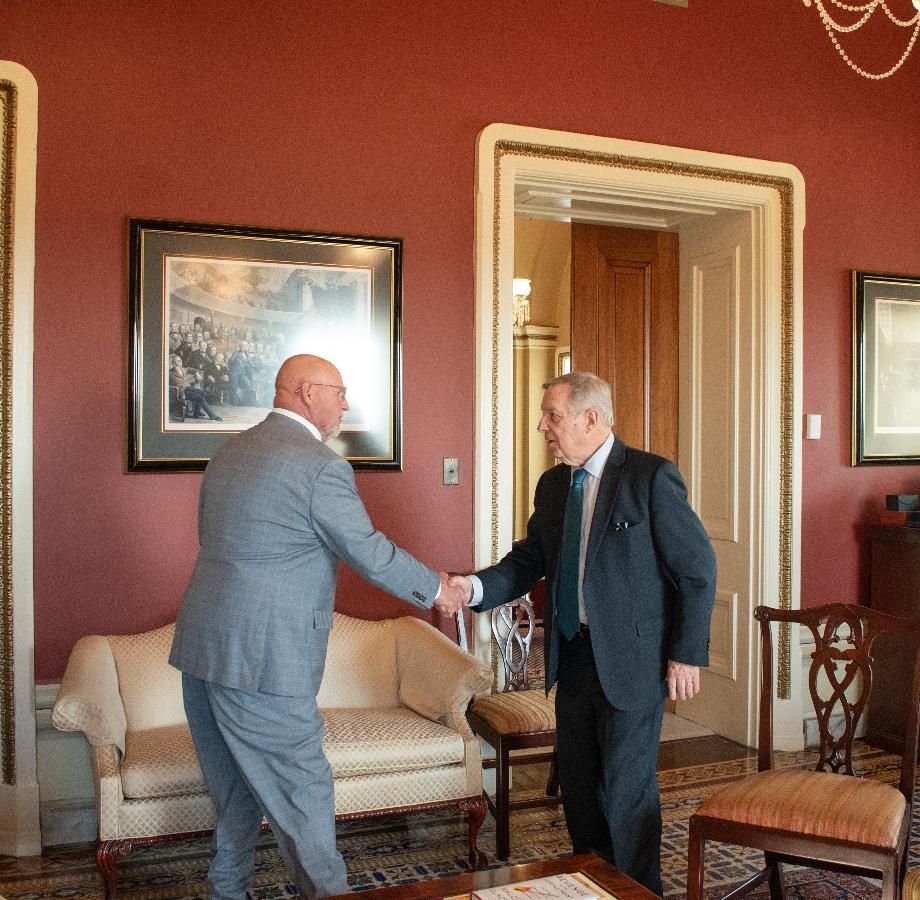 Two men in suits shaking hands in a room with red walls, a sofa, and artwork.