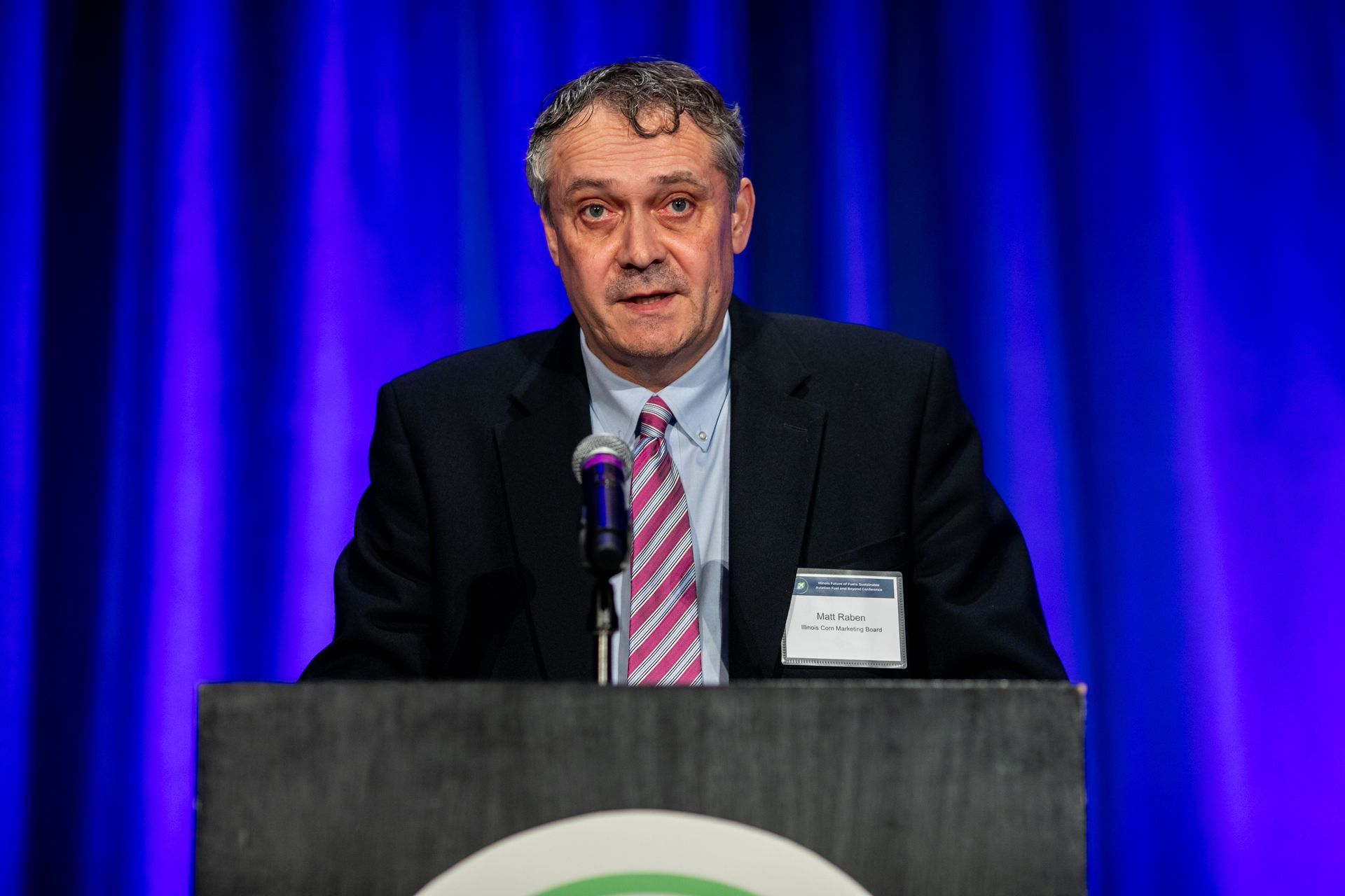 Man speaking at a podium, wearing a suit and tie, with a blue background.