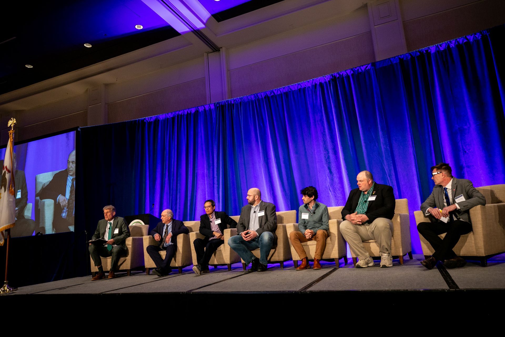 Panel of men sitting on stage at a conference, facing the audience, blue backdrop.