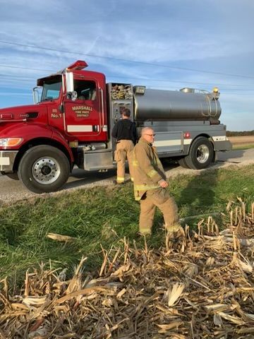 Firefighters near red truck with water tank in field, one examining debris.