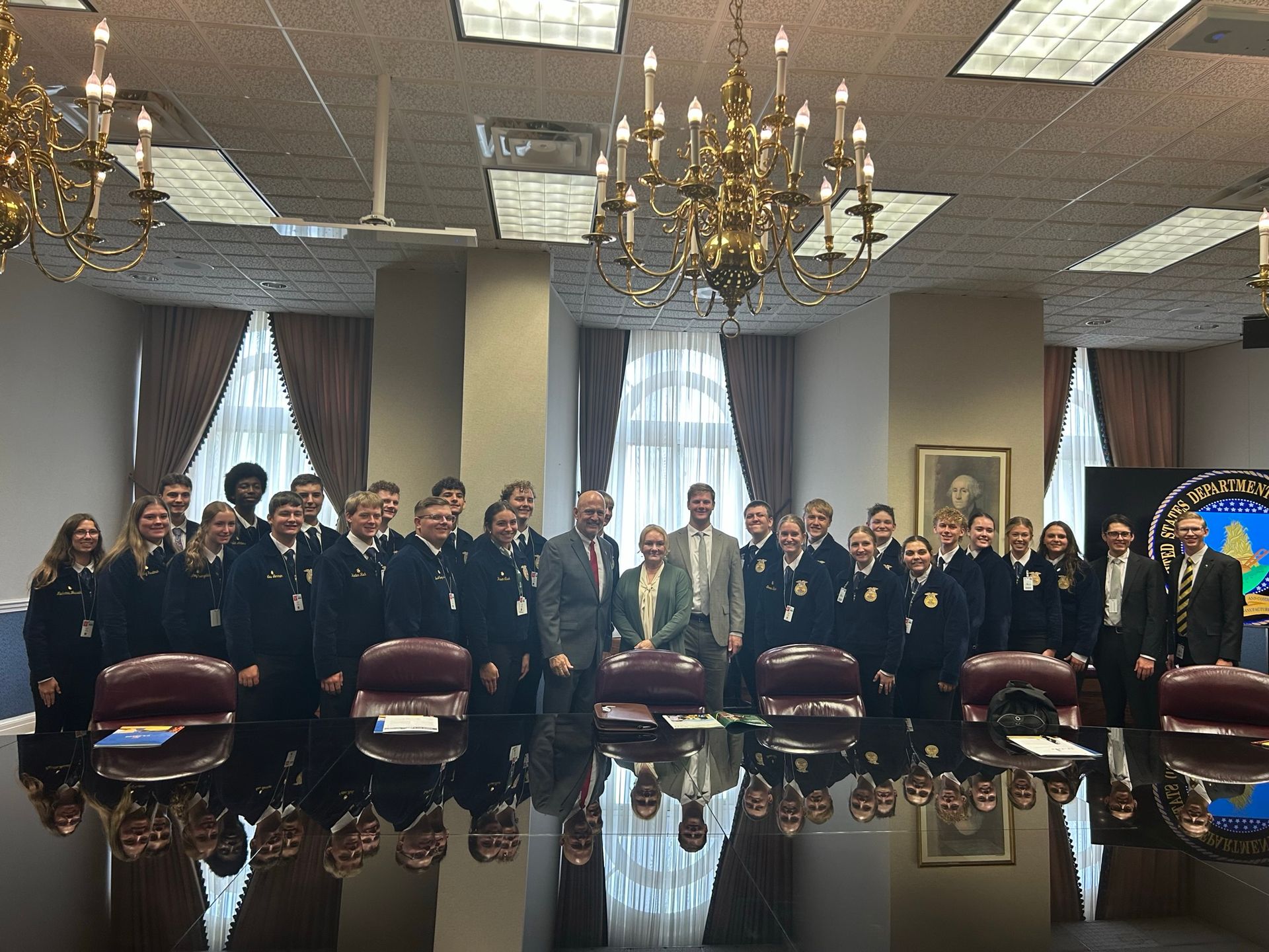 Group of students in blue jackets with adults, at a large table in a formal room.