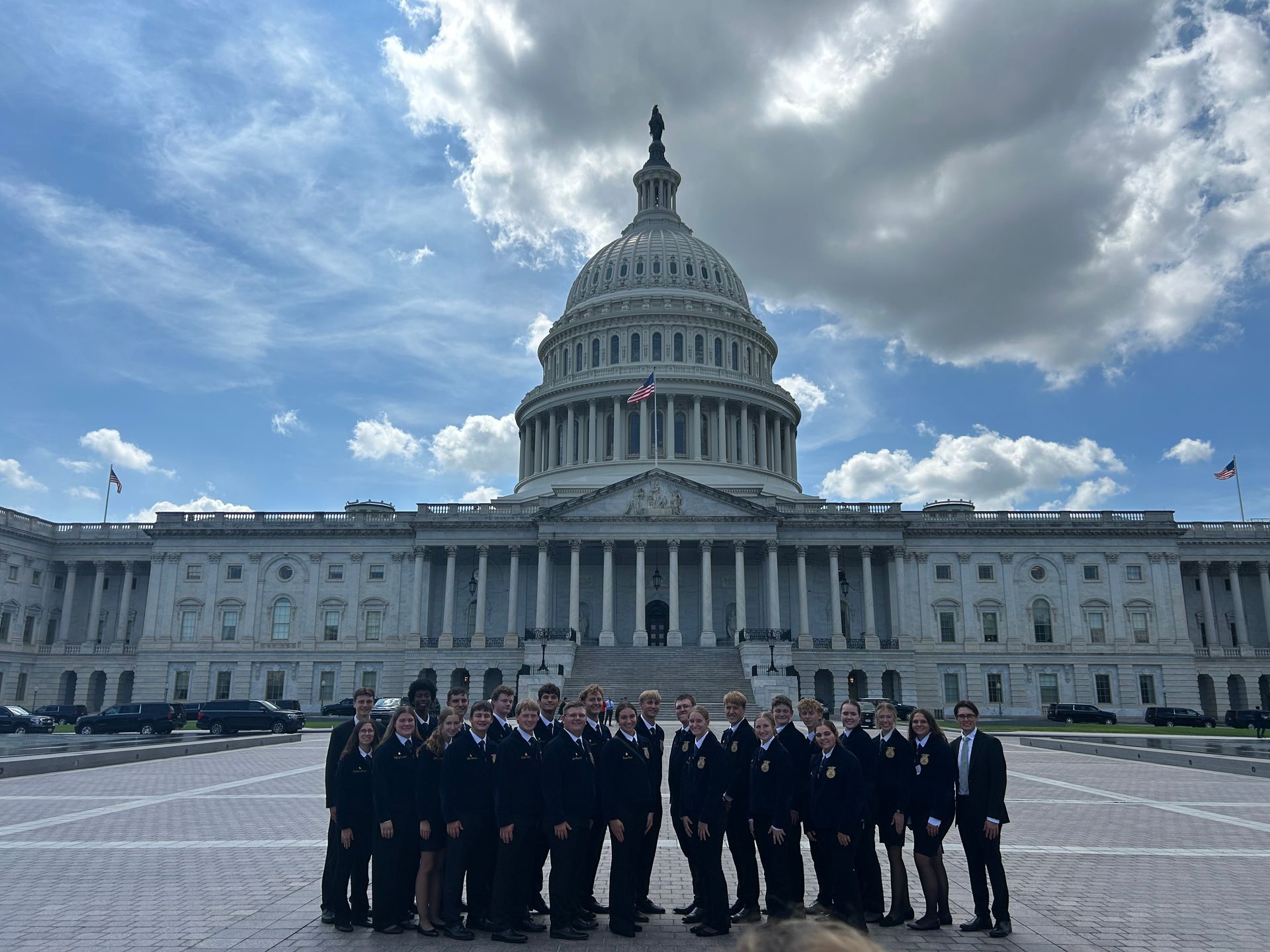 Group of people in dark suits in front of the US Capitol Building, under a cloudy sky.