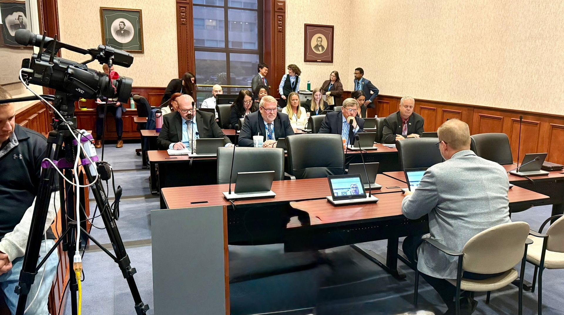 People seated at tables in a conference room, facing each other. Cameras and microphones are set up for a meeting.